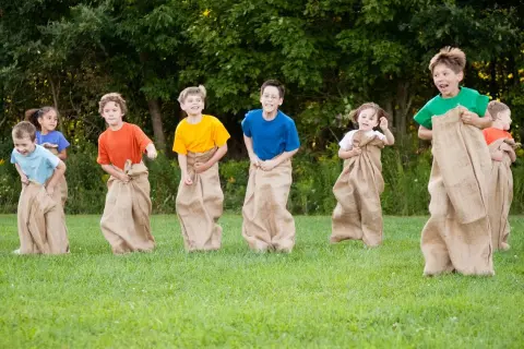 Barn hoppar säcklopp i en femkamp under midsommar. Glada barn i färgglada tröjor tävlar i säckar på en grön gräsmatta.