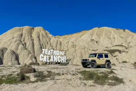 Un fuoristrada beige parcheggiato davanti al **teatro dei calanchi**, un paesaggio lunare con montagne erose e un cartello in legno.