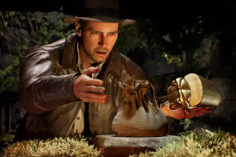 Indiana Jones i wielki krąg: archeolog z kapeluszem i skórzaną kurtką ostrożnie podnosi złoty artefakt w kształcie skarabeusza, obok leży skórzana sakiewka.