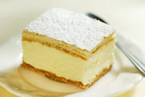 Pyszne ciasto budyniowe bez pieczenia, z kremowym nadzieniem i wafelkami, posypane cukrem pudrem.