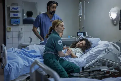 En ambulanssjuksköterska håller en patients hand medan en läkare ser på. Scen från en vaka serie.