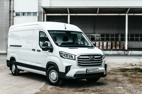 Biały Maxus Deliver 9 stoi przed magazynem. Ten wszechstronny van jest gotowy do pracy, przewożąc towary z miejsca na miejsce.