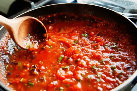 Gęsty, aromatyczny sos do spaghetti gotuje się na patelni. Drewniana łyżka miesza pomidorową masę z ziołami.