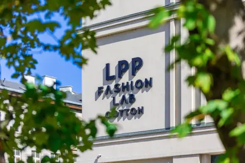 Napis LPP FASHION LAB COTTON na jasnym budynku, częściowo zasłonięty przez liście.