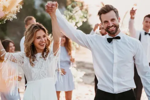 Glückliches Brautpaar feiert seine low budget hochzeit am Strand. Die Braut hält einen Blumenstrauß, der Bräutigam strahlt.