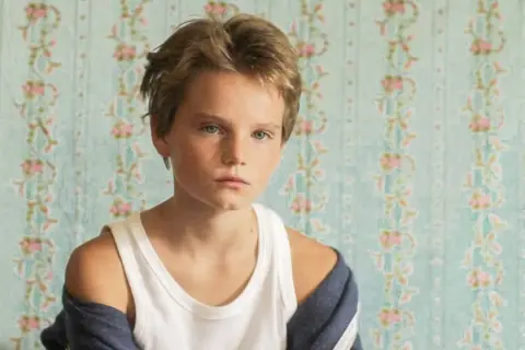 Un niño con cabello rubio y ojos azules, con una camiseta blanca y una chaqueta azul, parece un tomboy que es.