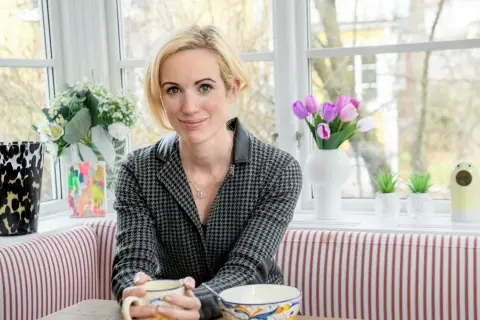 Jina Zachrisson sitter vid ett fönster med en mugg i handen, omgiven av blommor och konst.