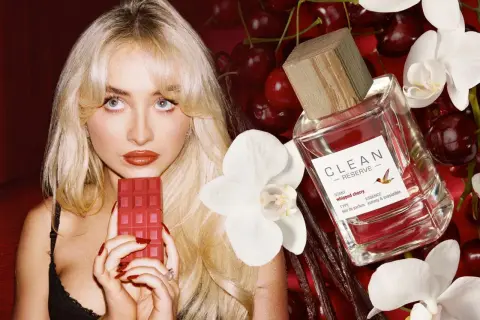 Kobieta z blond włosami trzyma czerwony batonik czekoladowy obok butelki perfum Clean Reserve Whipped Cherry, wiśni i białych kwiatów.