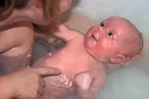Ein Baby genießt sein Bad, während eine Hand es sanft hält und eine andere auf seine Haut zeigt.