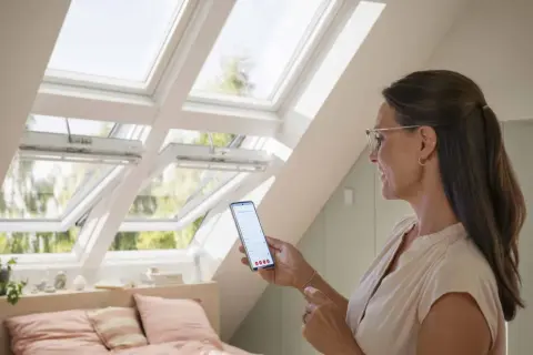 Une femme utilise son smartphone pour piloter des fenêtres de toit Velux.