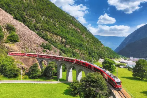 Czerwony pociąg przemierza malowniczy wiadukt w Alpach, na trasie Tirano - St. Moritz, otoczony zielonymi wzgórzami i błękitnym niebem.