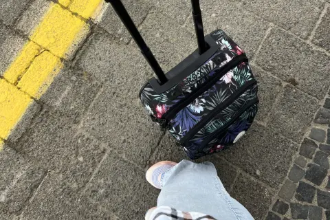 Kwiatowa walizka na kółkach i fragment nogi w jasnych jeansach na chodniku.