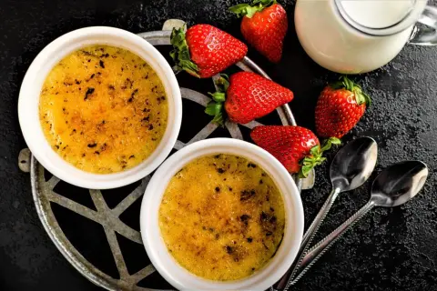 Zwei cremige Crème brûlée mit karamellisierter Kruste, garniert mit frischen Erdbeeren und einem Milchkännchen.