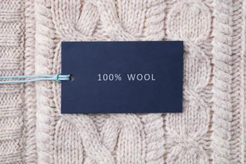 Metka z napisem "100% WOOL" na tle grubego, beżowego swetra w warkocze. To właśnie jest lambswool – czysta wełna.