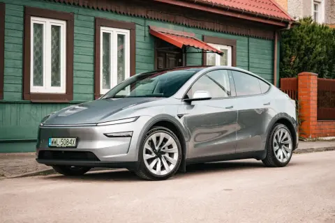Srebrny Tesla Y stoi przed zielonym domem. Zastanawiasz się, ile koni ma ten model?
