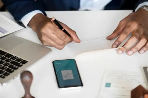 Uomo d'affari scrive su un libretto, con laptop e smartphone che mostrano grafici finanziari. Un esempio di come la categoria protetta art. 1 possa operare nel mondo del lavoro.