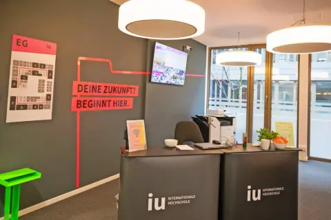 Hier ist die Rezeption der IU Internationale Hochschule. Ein Bildschirm zeigt Bilder, und ein Plan weist den Weg. Was ist die IU? Eine Hochschule, die deine Zukunft beginnt.