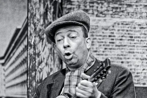 Stanisław Bard Starej Warszawy, w czapce i z gitarą, śpiewa na tle ceglanej ściany.