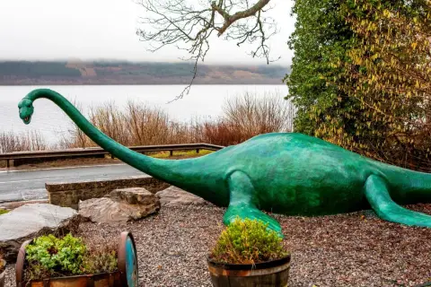 Zielony potwór z Loch Ness leży na kamieniach, z długą szyją wyciągniętą w stronę jeziora.