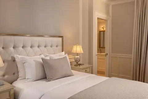 Elegancka sypialnia z dużym łóżkiem, pikowanym zagłówkiem i stylowymi lampkami nocnymi. Widok na łazienkę i zasłony. Idealne miejsce dla wynajmującego szukającego komfortu.