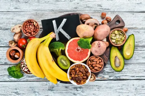 Frutta e verdura che mostrano dove si trova la vitamina K2: banane, broccoli, avocado, pomodori, uva passa, mandorle, semi di zucca, patate, funghi, nocciole.