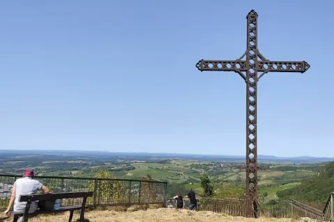 La croix du Dan domine un paysage verdoyant. Deux personnes profitent de la vue depuis un banc.