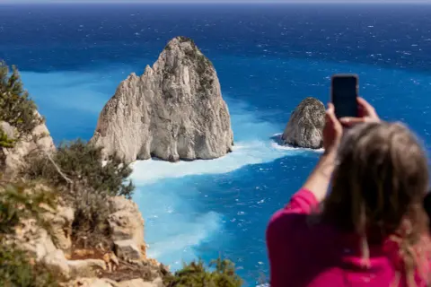 Widok na turkusowe morze i skaliste wysepki. Kobieta robi zdjęcie telefonem, uwieczniając piękno Zakynthos. Idealne miejsce na wycieczki fakultatywne.