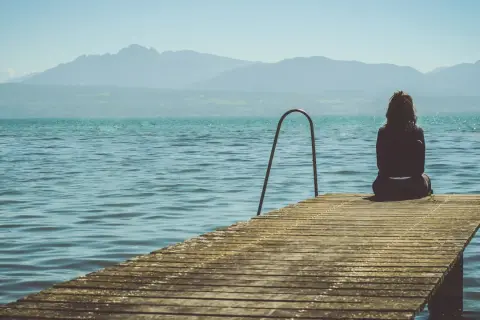 Mujer sentada en un muelle, contemplando el lago y las montañas. Pensamientos de frases de preocuparse por alguien flotan en el aire.