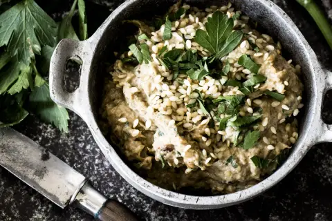 Cremiges Baba Ganoush, garniert mit Sesam und Petersilie, in einem rustikalen Topf. Daneben ein Messer und frische Kräuter.