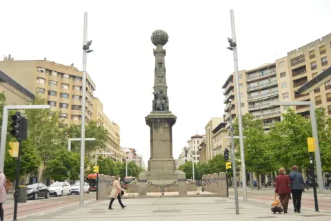 Monumento en la plaza Aragón con edificios modernos y árboles verdes. Dos mujeres caminan con equipaje.