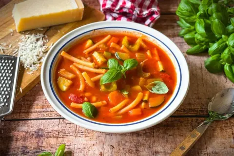 Herzhaftes rezept minestrone original mit viel Gemüse und Pasta, garniert mit frischem Basilikum. Daneben Parmesan und ein Löffel.