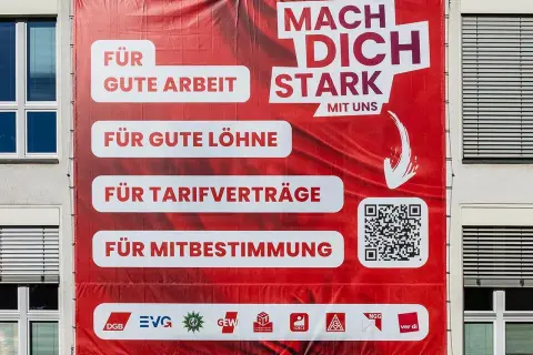 Großes rotes Banner mit der Aufschrift "Mach dich stark mit uns". Es wirbt für gute Arbeit, Löhne, Tarifverträge und Mitbestimmung. Was ist ver.di? Die Antwort steht unten.