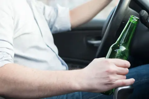 Mężczyzna za kierownicą trzyma butelkę piwa. Nie warto wiedzieć, ile % ma piwo, gdy prowadzisz.
