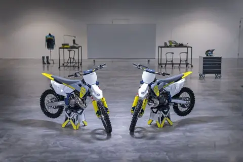 Dwa motocykle crossowe Husqvarna TC 125 w białym i żółtym kolorze stoją na stojakach.