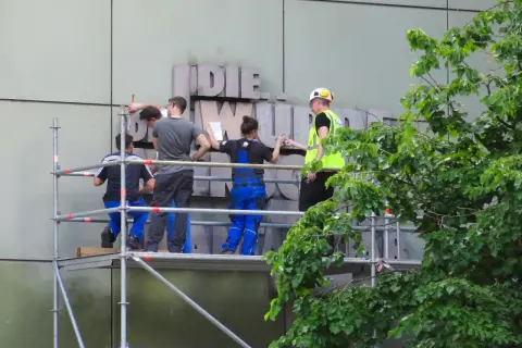 Arbeiter auf Gerüst montieren Buchstaben an einer Fassade. Ein Mensch in Warnweste gibt Anweisungen.