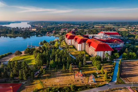 Hotel Gołębiewski w Mikołajkach, z czerwonymi dachami, otoczony zielenią i jeziorem, zaprasza na wypoczynek. Ile lat ma hotel? To miejsce z historią!