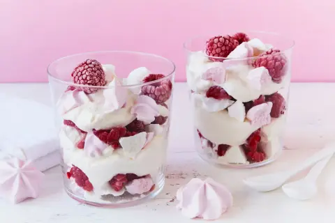 Zwei Gläser mit Himbeer Baiser Dessert, geschichtet mit Sahne, frischen Himbeeren und Baiserstückchen.
