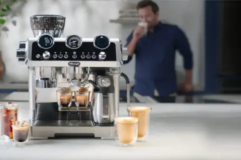 Macchina per caffè De'Longhi in azione, con chicchi pronti per la produzione del caffè. Un uomo assapora una bevanda calda.