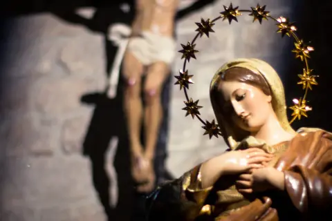 Figurka Matki Bożej z gwiazdami na głowie, w tle ukrzyżowany Jezus. Symbol bólu i cierpienia, jak w różańcu części bolesne.