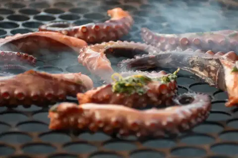 Tentáculos de pulpo a la brasa humeantes sobre una parrilla. El pulpo se cocina lentamente, adquiriendo un color dorado y un aroma irresistible.
