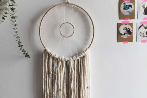 Un attrape-rêves fait main, parfait pour un tuto attrape rêve. Des fils de laine beiges et gris tombent, ornés de perles en bois.
