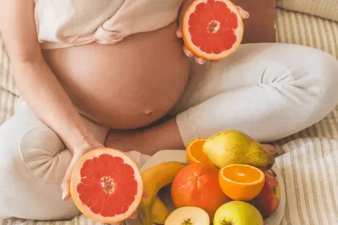 Ciężarna kobieta trzyma połówki grejpfruta, obok talerz z owocami. Czy w ciąży można jeść pomarańcze? Tak, to zdrowe!
