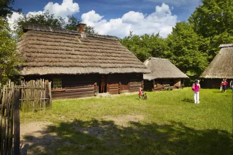 Drewniane chaty ze słomianymi dachami, trawiasty dziedziniec i ludzie.