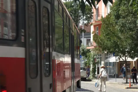 Przejeżdżający tramwaj w słoneczny dzień w Sofii. Kobieta w kapeluszu idzie ulicą z torbami.