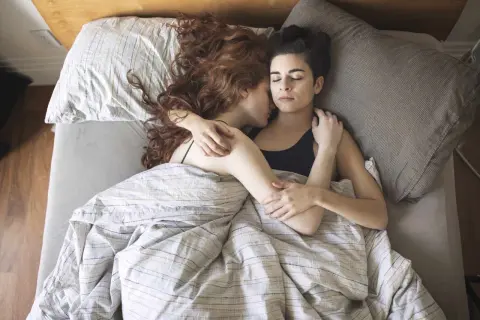 Two women sleeping together, embracing warmly under a striped duvet.