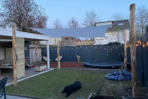 Sonnensegel befestigen Ideen: Ein graues Sonnensegel spannt sich über einen Garten mit Trampolin und Sitzbereich. Holzpfosten dienen als Befestigungspunkte.