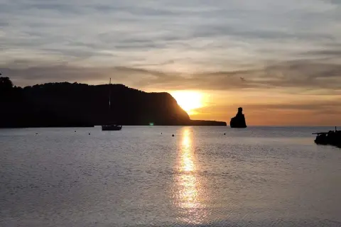 Un velero navega en la serena bahía mientras el sol se pone en Benirràs, tiñendo el cielo de tonos dorados y naranjas.