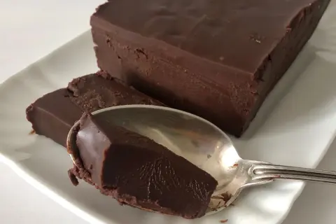Una mattonella di cioccolato fondente, densa e lucida, su un piatto bianco. Un cucchiaino ne ha prelevato un pezzo.