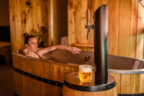 Entspannende Frau in einem Bierfass-Bad, daneben ein frisch gezapftes Pils. Ein einzigartiges Bier-Spa-Erlebnis in Pilsen.