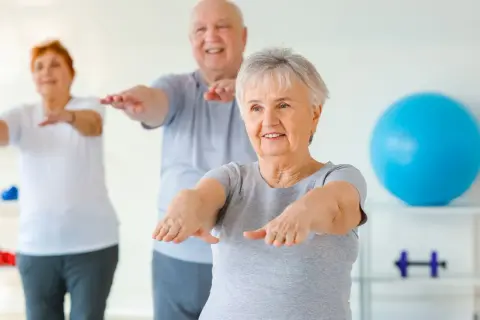 Trois seniors souriants pratiquent la gym chinoise pour seniors, bras tendus, dans une salle lumineuse.